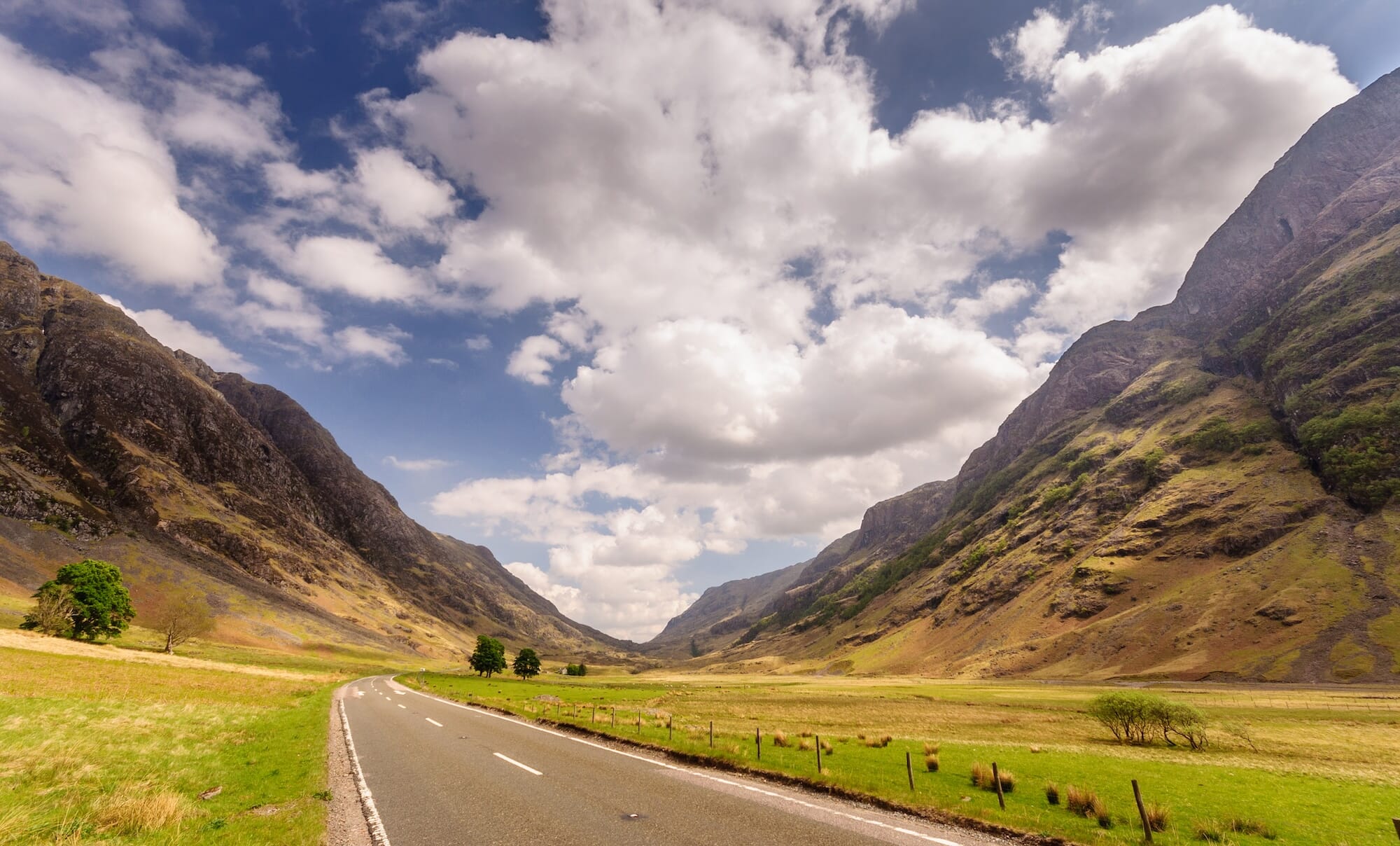 The A82 road crosses pasture fields in the valley floor of Glen Coe, under the mountains of the West Highlands of Scotland.